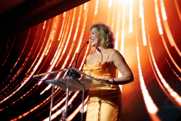 A woman in an orange dress delivers a speech at a podium during the SBID Awards